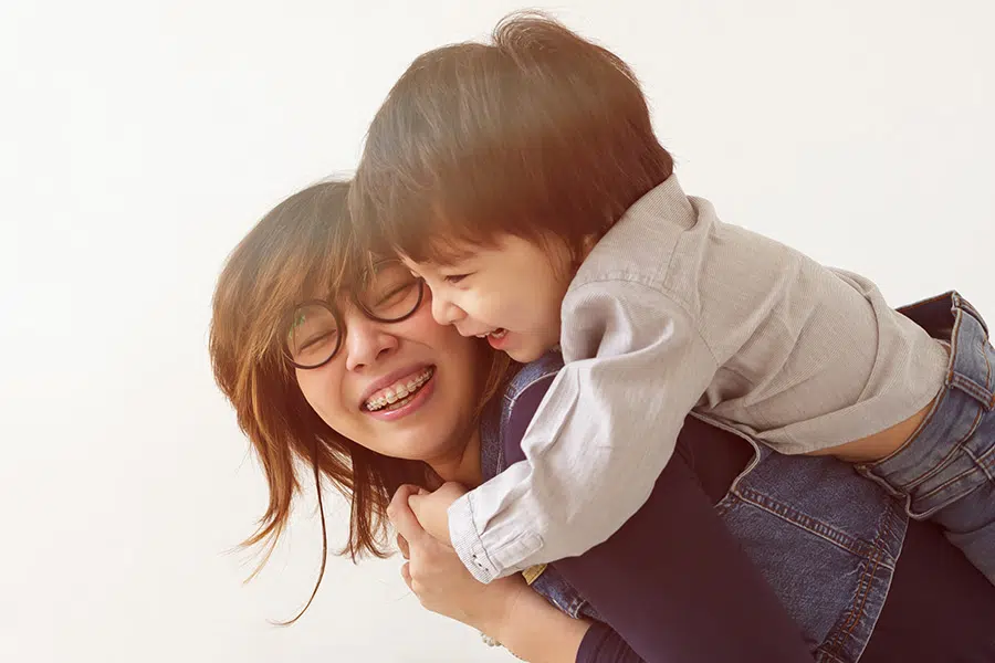 An adult female wearing metal braces and laughing with her young son on piggyback
